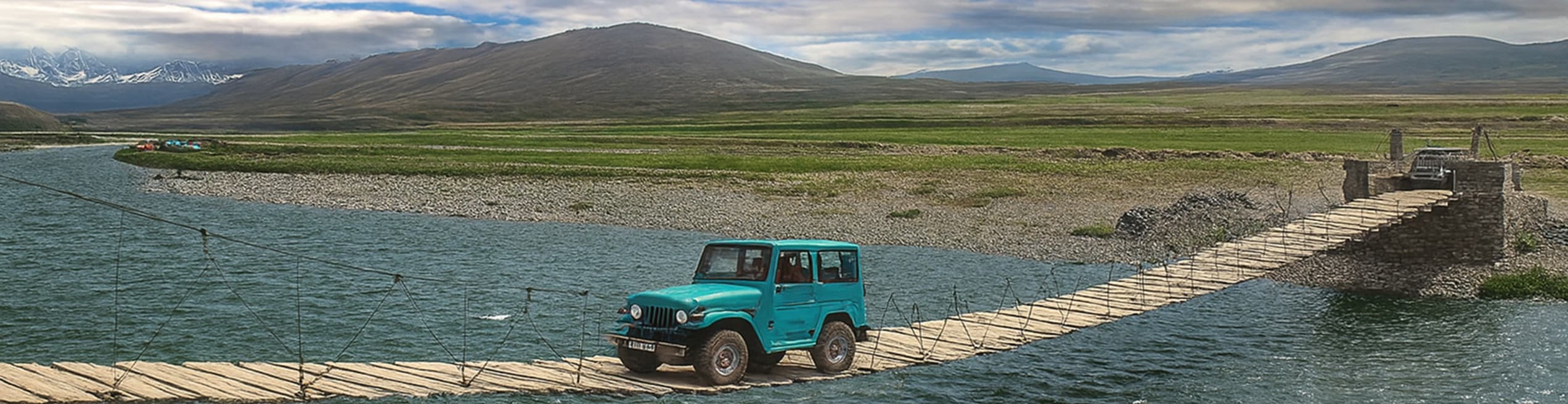 A jeep driving across a wooden bridge over a river in Deosai Plains, surrounded by vast grasslands and mountains.
